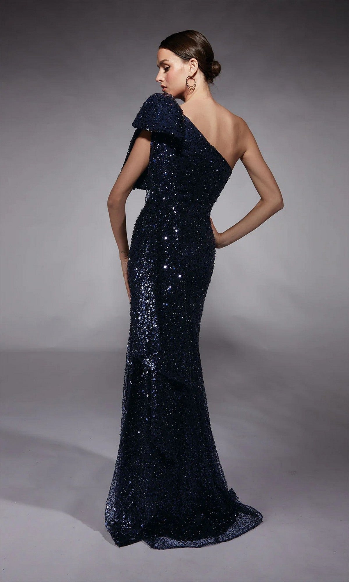 A woman models the Alyce 27771 long formal dress, a dark blue one-shoulder sequin evening gown, posing with her back slightly turned against a neutral background.